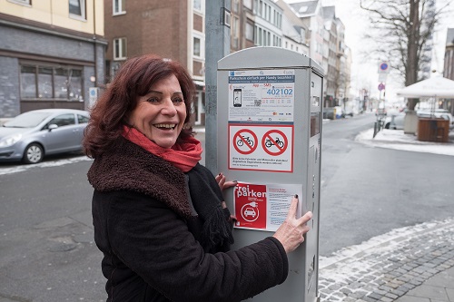 Umweltdezernentin Helga Stulgies bringt am Parkscheinautomaten einen Aufkleber an, der auf das Angebot des freien Parkens für registrierte Elektrofahrzeuge hinweist.